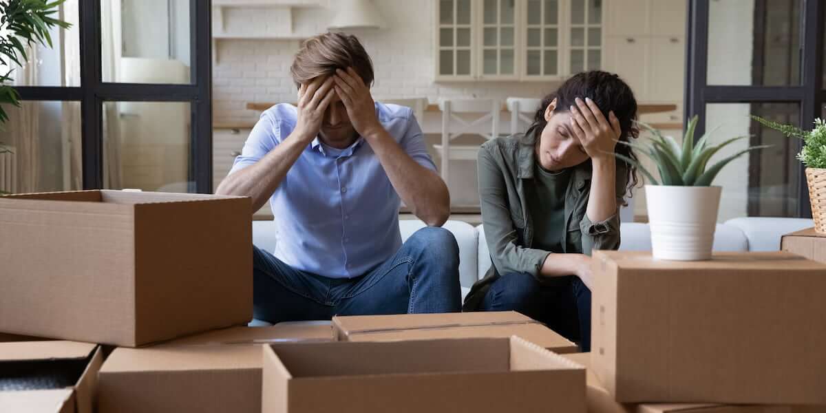 A stressed couple sitting among moving boxes, holding their heads in frustration, illustrating the challenge of moving and the importance of learning how to make moving less stressful.